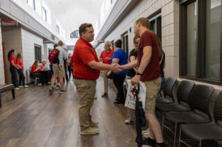 Two men shake hands in a hallway lined with chairs, perhaps after discussing scholarships or financial aid opportunities. Several people are in the background, some sitting and some standing, wearing casual clothes. One man holds a bag and a rolled-up poster. The setting appears to be a campus or office.