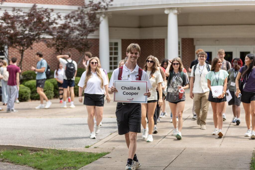 A group of students walks outside a building. A person at the front holds a sign reading “Chaille & Cooper.” Most wear casual clothing and carry bags or drinks. Trees and a brick building are visible in the background.