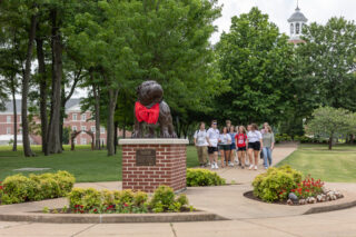 A group of people walk on a campus pathway near a statue of a bulldog wearing a red scarf. The area is landscaped with flowers, trees, and brick buildings, including one with a white clock tower in the background.