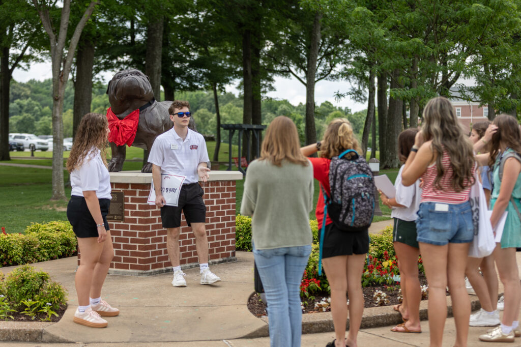 A tour group stands near a dog statue with a red cloth around its neck. The guide, wearing sunglasses and a white shirt, speaks to the group. Trees and parked cars are visible in the background.