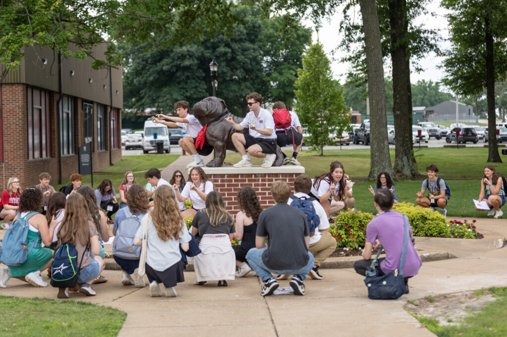 A group of students gathers outdoors around a brick structure, with a few standing or crouching on top, while others sit or kneel on the pavement and grass, listening. Trees and buildings are visible in the background.