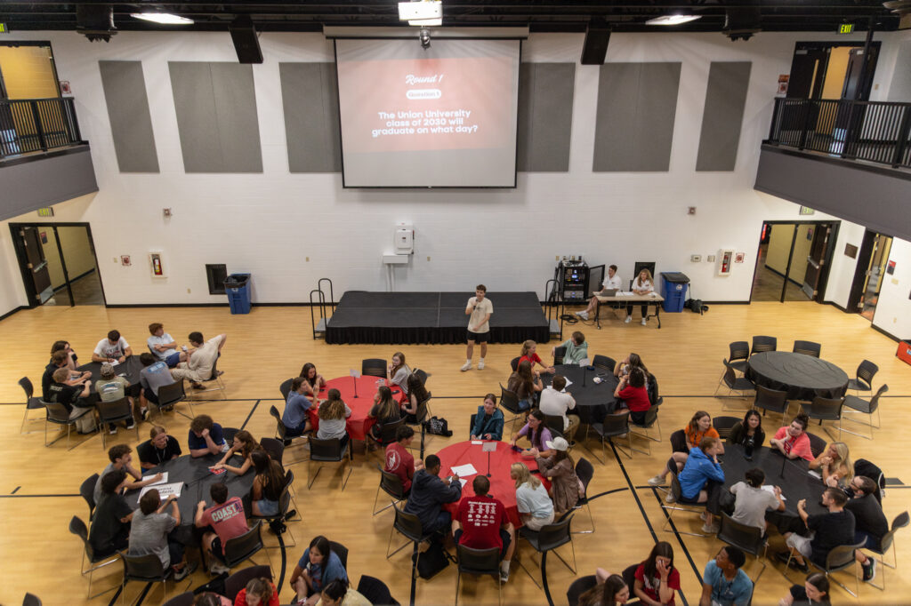 A group of people sit at round tables in a gymnasium, facing a large screen showing a question about Union University graduation dates. One person, possibly from Residence Life, stands at the front with a microphone.