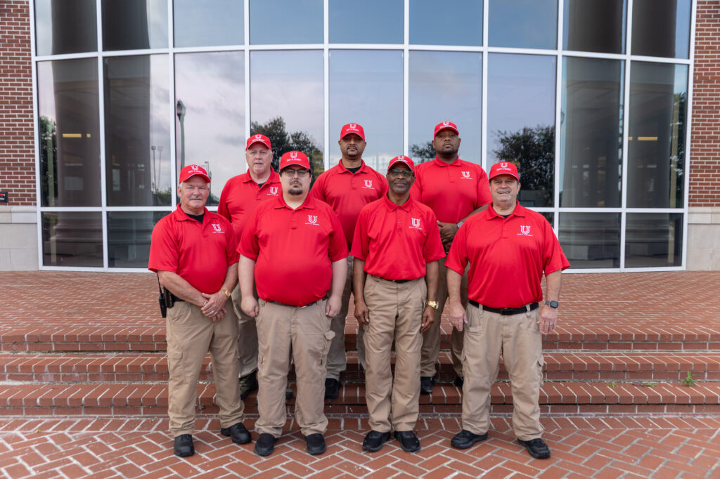 Seven men stand on brick steps in front of a building with large windows, all wearing red shirts, khaki pants, and red caps. The group is posing for a photo, and the background shows the building’s glass facade.