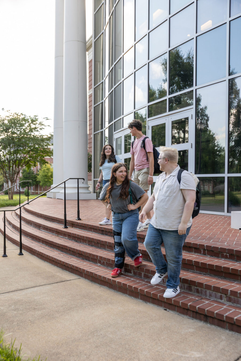 Four students with backpacks walk down the steps outside a modern building with large glass windows and brickwork. They appear to be engaged in conversation and are casually dressed.