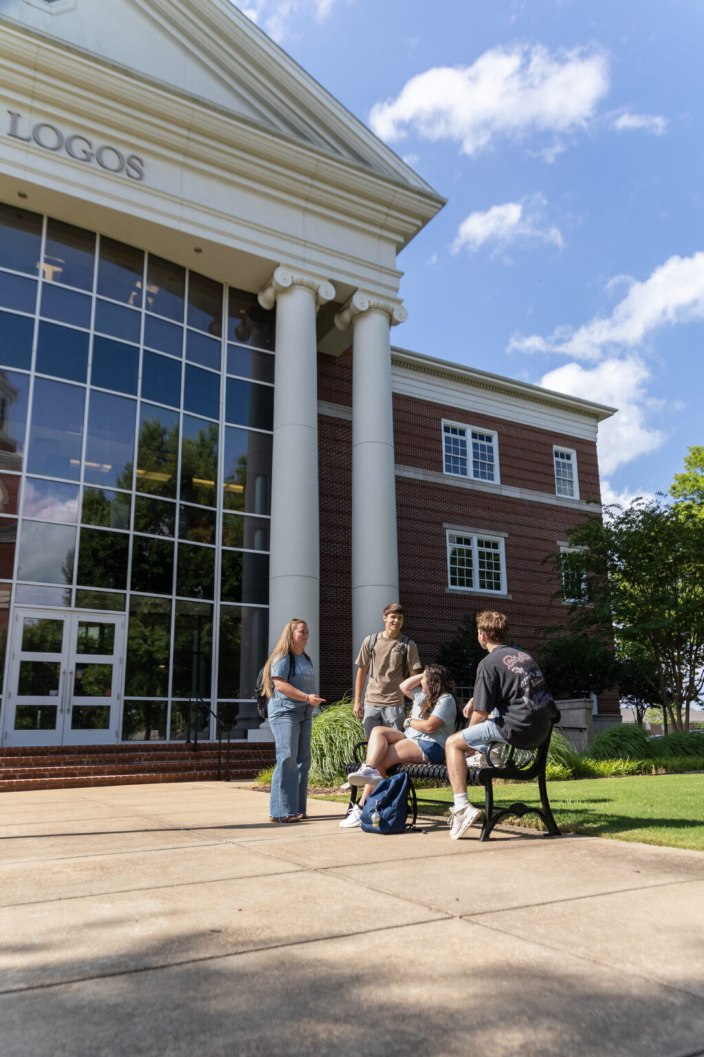 Four young people gather outside a large building with tall white columns and large windows. Two are standing and two are sitting on a bench, with backpacks nearby. The sky is clear with a few clouds.