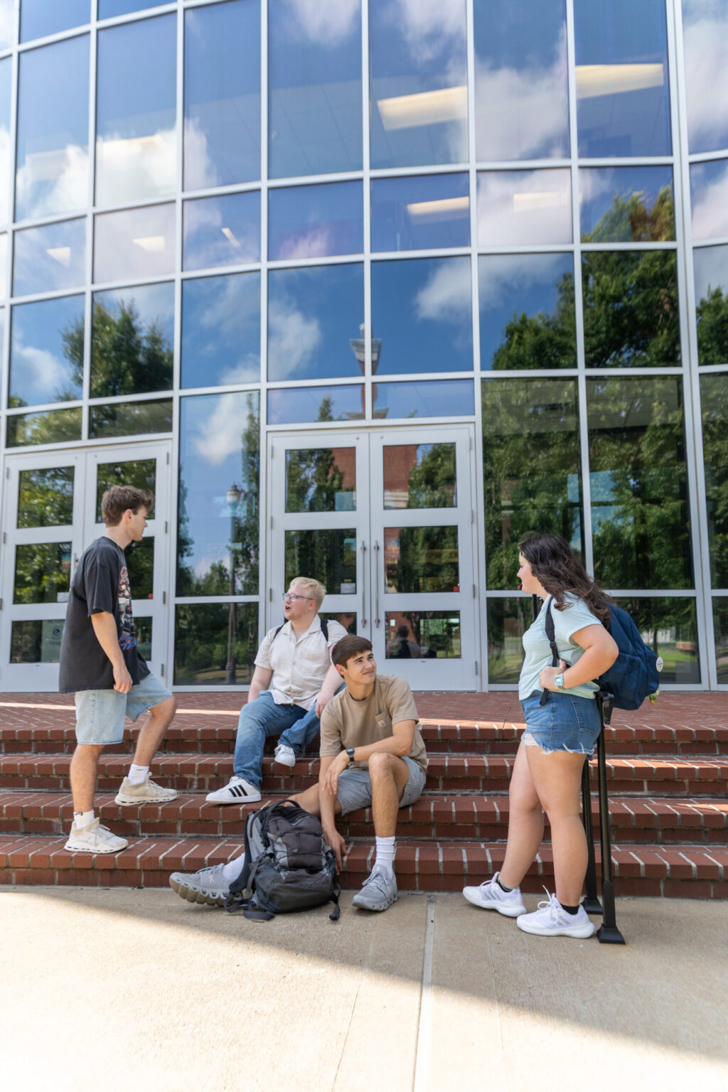 Four students with backpacks gather and talk on brick steps outside a modern building with large glass windows reflecting the sky and trees. Two students sit while two others stand.