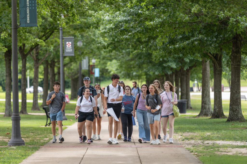 A group of students walk together on a tree-lined sidewalk during the day, carrying backpacks and talking. Some are smiling. Campus signs and green grass are visible in the background.
