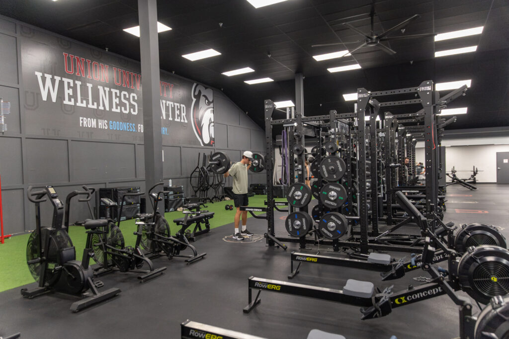A person lifts a barbell in a spacious gym with weight racks, rowing machines, and exercise bikes. The wall displays “Union University Wellness Center” with a bulldog logo. The gym is well-lit and clean.