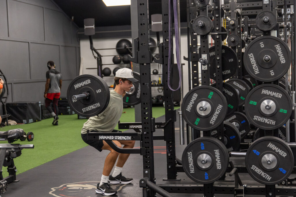 A person wearing a white hat squats with a barbell on their shoulders in a gym. Weight plates are stacked on racks nearby. Other gym equipment and people are visible in the background.