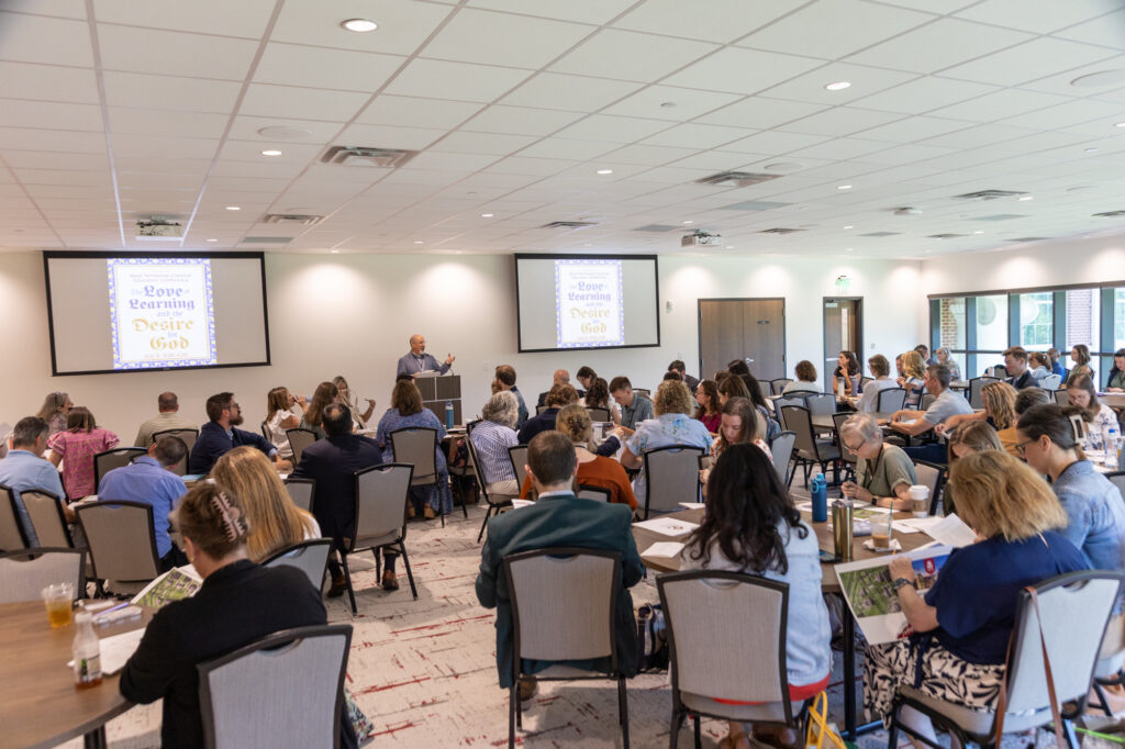 A group of people sit at tables in a conference room, listening to a speaker at a podium. Presentation slides read “Love Learning Desire God.” Members of the Honors Community have papers, drinks, and notebooks on the tables.