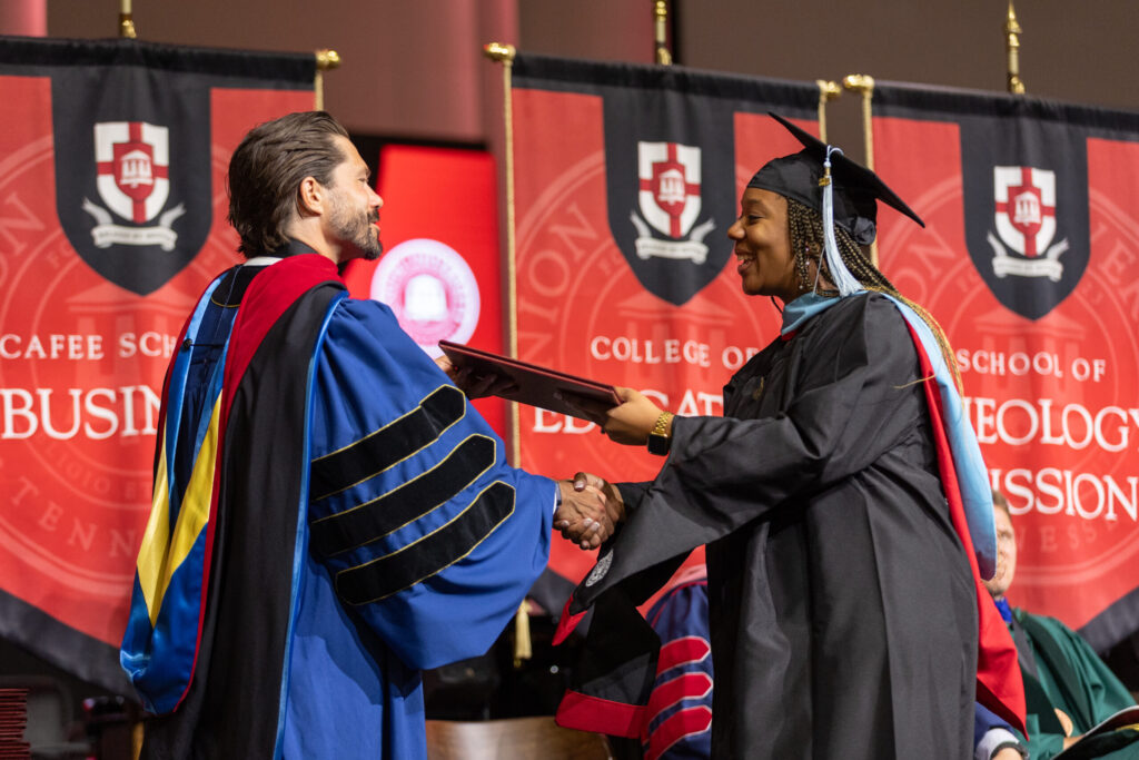 A graduate in cap and gown shakes hands with a faculty member while receiving a diploma on stage, symbolizing the proud tradition of Union graduates. Red banners with academic symbols and text are displayed in the background, echoing the belief that to whom much is given, much is required.