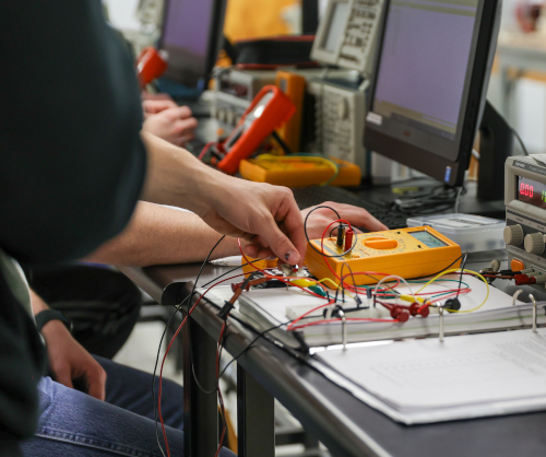 A Union engineering student uses a yellow multimeter to test an electronic circuit on a desk, with wires, electronic components, computers, and equipment visible in the background—demonstrating practical skills that contribute to the high FE exam pass rate.