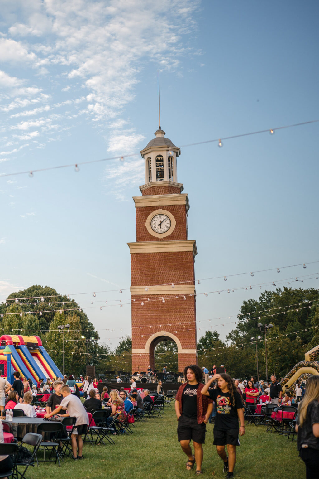 People gather at outdoor tables on a grassy lawn with a tall, red-brick clock tower in the background. Two people walk in the foreground; an inflatable slide and ride can be seen to the left. String lights hang overhead.