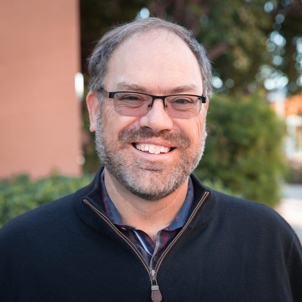 A man with glasses and a beard, wearing a black zip-up sweater over a plaid shirt, smiles outdoors in front of greenery and a building.