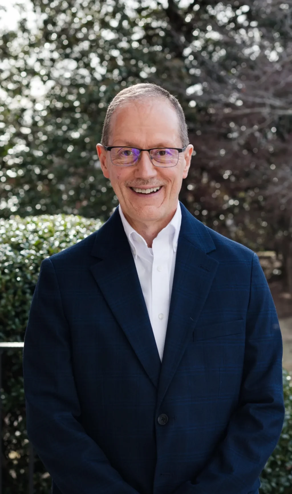 A man wearing glasses, a dark blue blazer, and a white shirt is standing outdoors in front of greenery and trees, smiling at the camera.