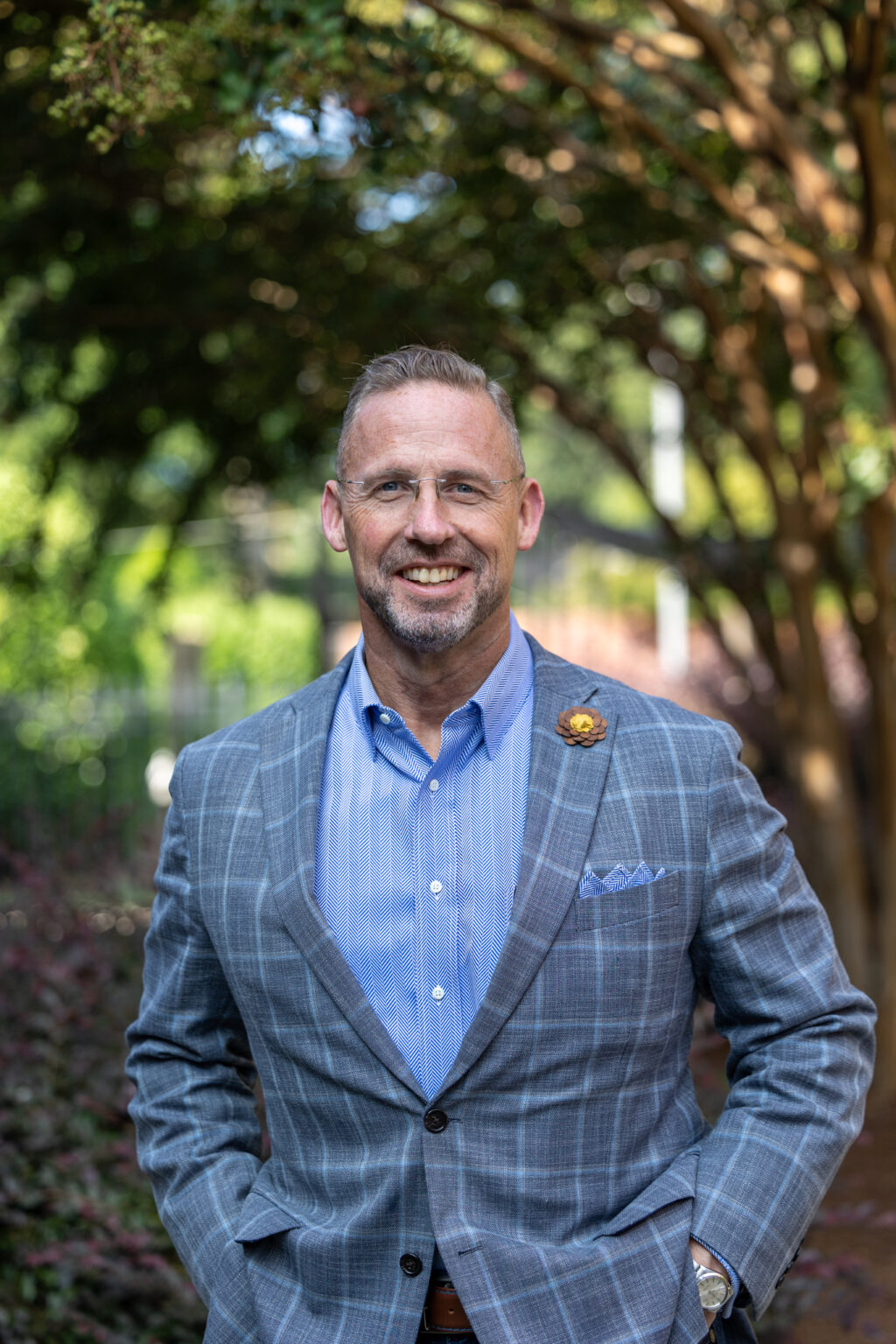 A man with short hair, glasses, and a trimmed beard is standing outdoors, wearing a gray plaid suit jacket, blue shirt, and floral lapel pin. Trees and greenery are visible in the background.