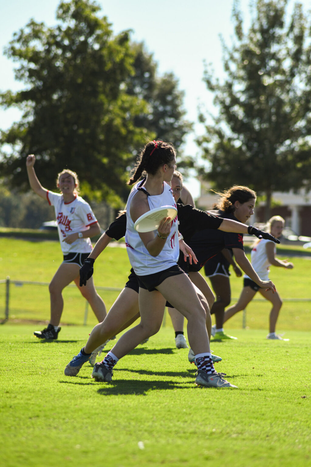 A woman in a white jersey prepares to throw a frisbee during an outdoor ultimate frisbee game. Other players in black and white jerseys are running on the grassy field under a sunny sky.
