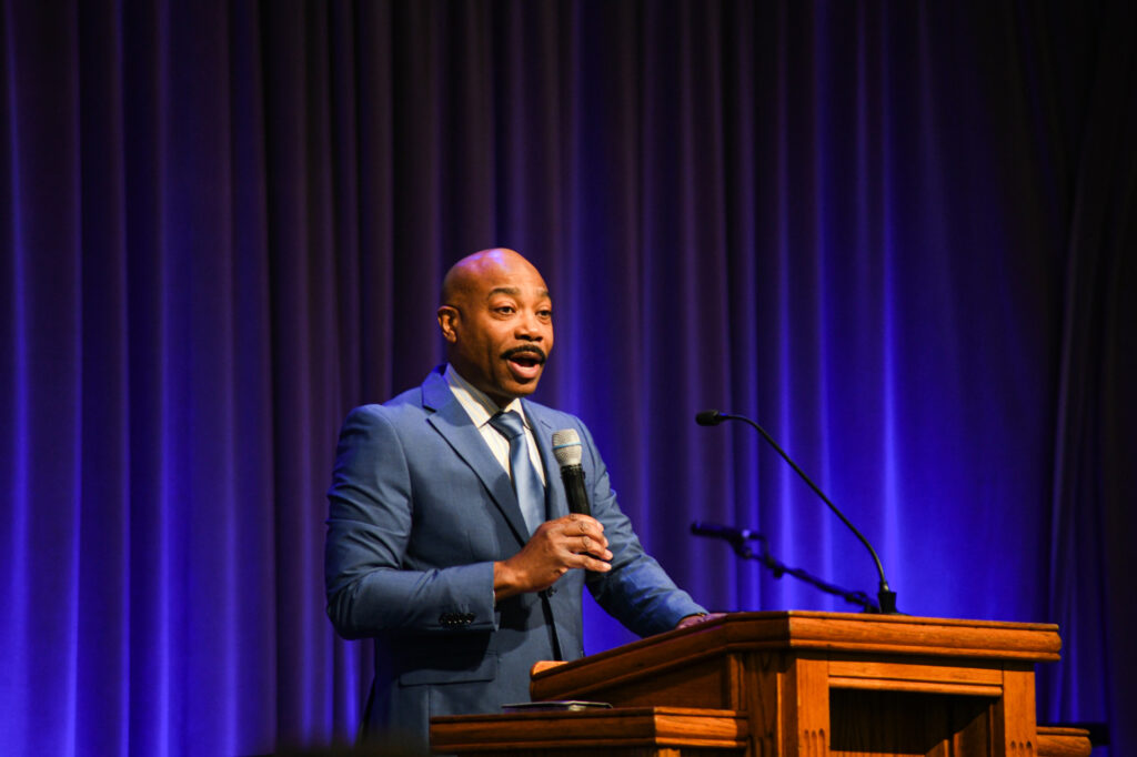 A man in a blue suit speaks into a microphone at a wooden podium. He is standing in front of a purple curtain and holding a piece of paper, with an additional microphone visible on the podium.