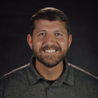 A man with short brown hair and a beard is smiling at the camera. He is wearing a dark grey collared shirt and is posed in front of a dark, plain background, ready for the Equip 2026 Conference.