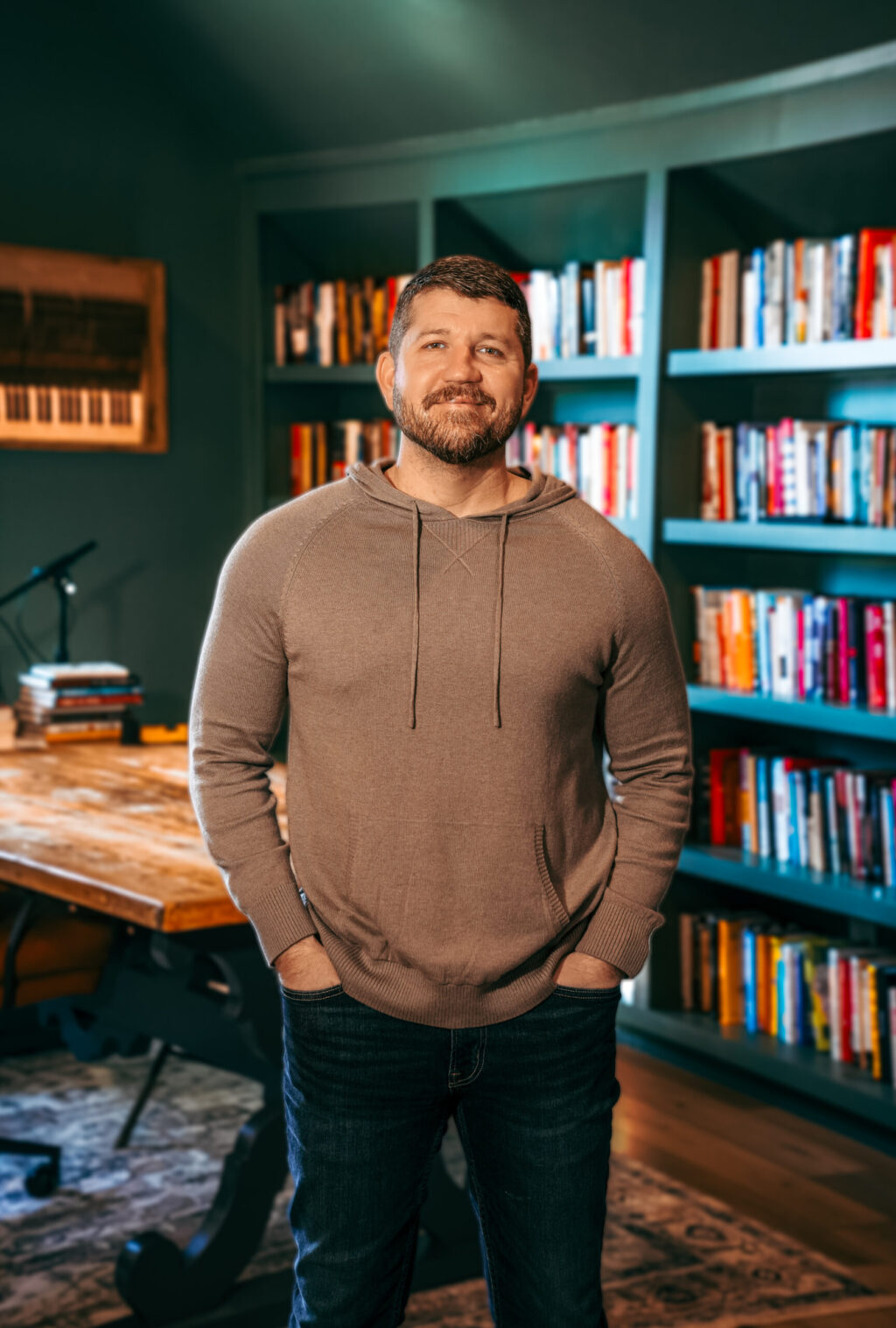 A man with short hair and a beard stands in front of a wooden desk and bookshelves filled with books in a home office or library setting. He is wearing a tan hoodie and jeans, with his hands in his pockets.