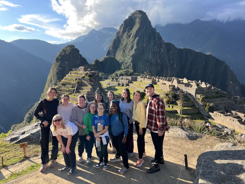 A group of people pose for a photo in front of the ancient ruins of Machu Picchu in Peru, with terraced stone structures and mountains visible in the background under a partly cloudy sky.