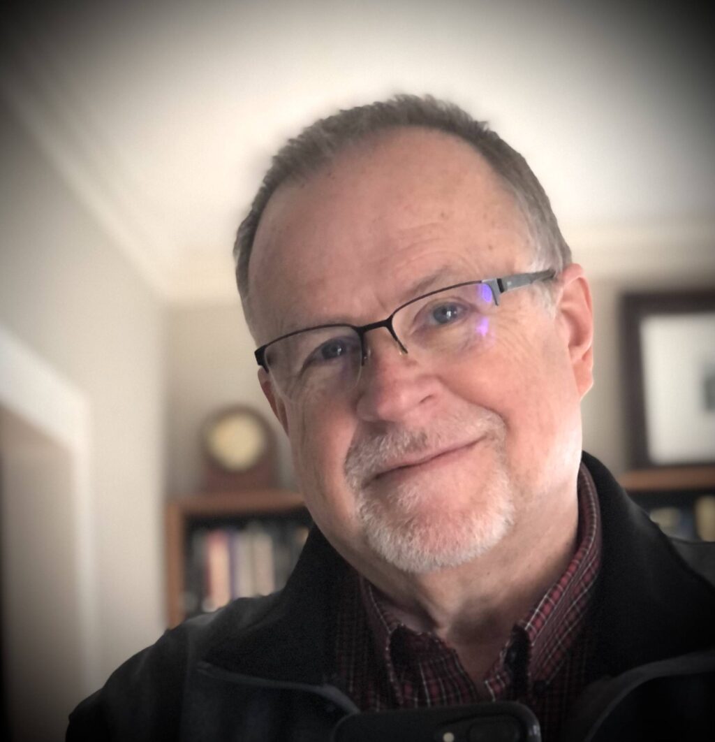 A middle-aged man with glasses, short gray hair, and a trimmed beard smiles at the camera. He is wearing a dark jacket over a checkered shirt, with a blurred bookshelf and framed picture in the background.
