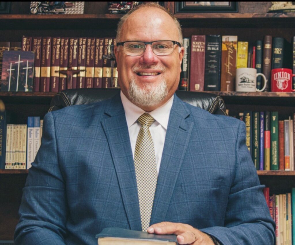 A man with glasses and a short white beard is sitting in front of a bookshelf filled with books. He is wearing a blue suit and a yellow patterned tie, holding a book with both hands. The serene setting hints at the quiet reflection often found in chapel moments, making it easy to imagine Trent Bullock preparing for an inspiring address.