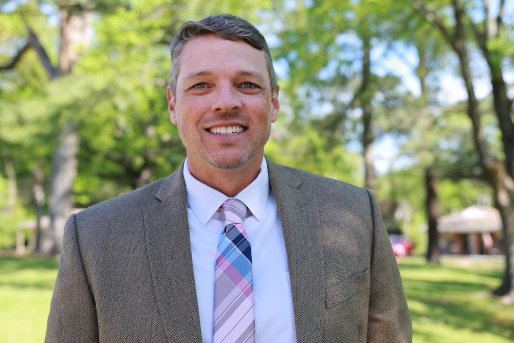 A man wearing a brown suit, white shirt, and multicolored striped tie stands outdoors in a park-like setting with green trees and sunlight in the background.