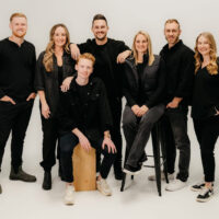 Seven adults pose in a studio with a white background. Six stand and one sits on a wooden box. Dressed in black or dark clothing, they smile warmly at the camera—ready to represent Equip 2026 at this year’s Equip Conference.