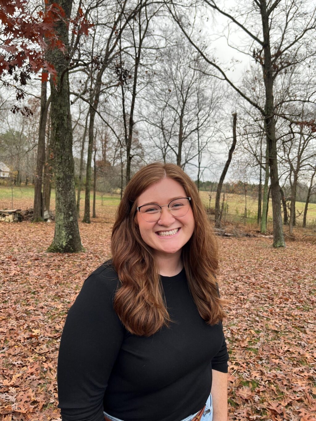 Juliana White, a woman with long brown hair and glasses, smiles outdoors in a wooded area with fallen leaves on the ground. The background reveals bare trees and a grassy field under a cloudy sky, capturing the tranquil beauty near the chapel.