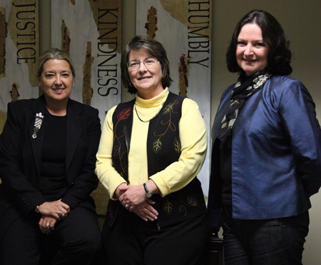 Three women stand indoors in front of decorative banners that read Justice, Kindness, and Humbly. Dressed in business-casual attire, they represent the Center for Just Communities and look toward the camera with purpose.