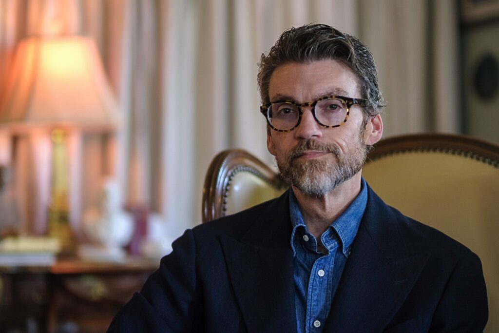 A man with gray hair, a beard, and glasses sits in a tan upholstered chair. He is wearing a blue shirt with a dark blazer. A lamp and curtains are visible in the softly lit background.