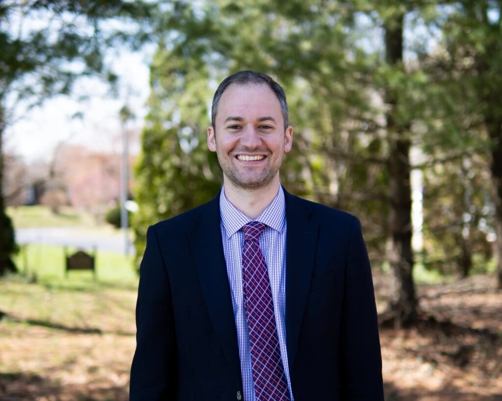 A man in a dark suit, light shirt, and patterned tie stands outside, smiling at the camera. Trees and greenery are in the background on a sunny day.