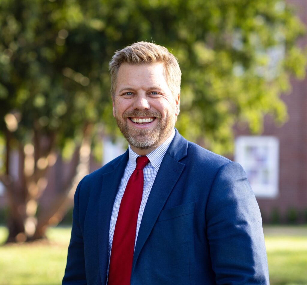 A man in a blue suit and red tie stands outdoors, smiling at the camera. Green trees and a brick chapel building are visible in the blurred background, suggesting Stephen Presley is enjoying a pleasant day outside.