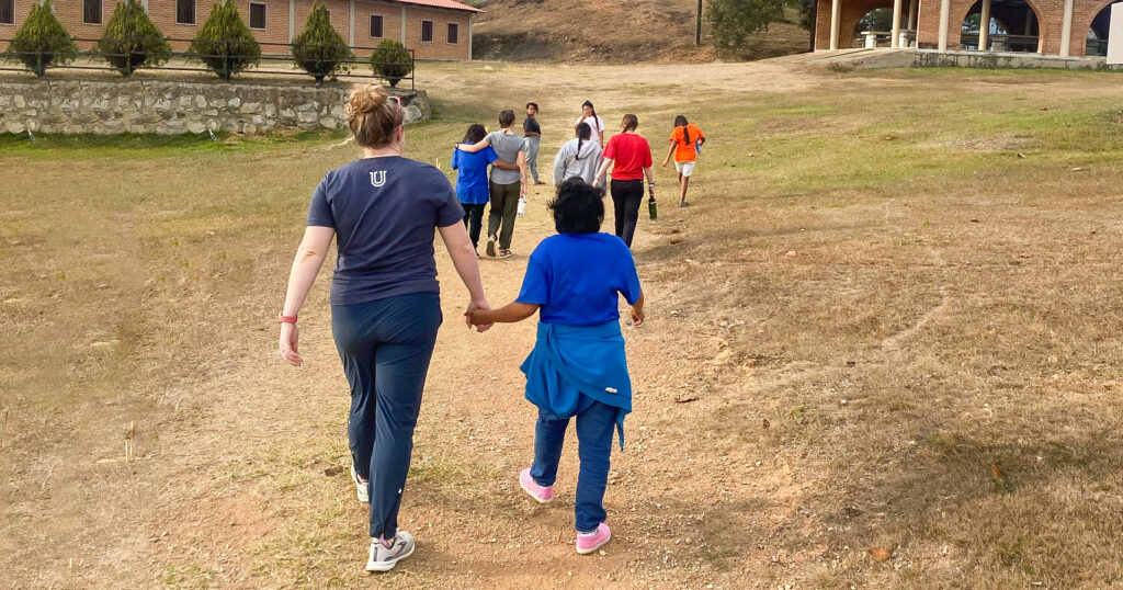 A group of people walk uphill on a dirt path in a grassy area, with two people in the foreground holding hands. Several buildings with brick walls are visible in the background.