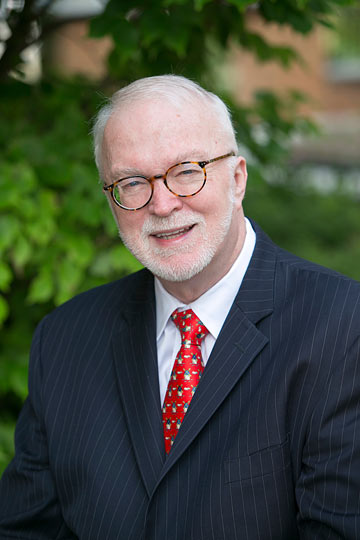 An older man with white hair, glasses, and a white beard is smiling. He is wearing a dark pinstripe suit, a white shirt, and a red tie with a small pattern. Green foliage and the blurred outline of a chapel can be seen in the background. This distinguished gentleman is Trent Bullock.