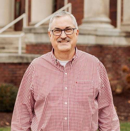 A man with gray hair and glasses stands outside in front of a brick building, wearing a red and white checkered button-up shirt and smiling at the camera—this is Trent Bullock, posed in front of the chapel’s entrance.