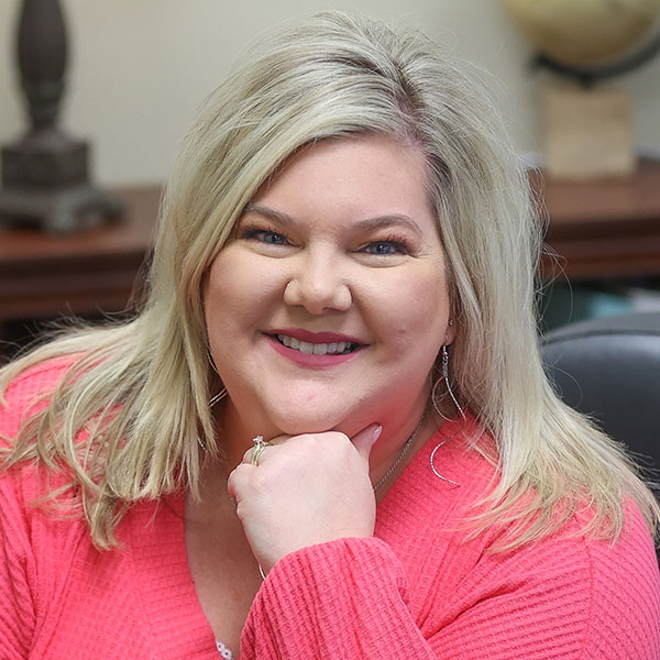 Jamie Connor, a woman with long blonde hair and a pink top, sits at a desk, resting her chin on her hand and smiling at the camera. The background includes a lamp and a globe.