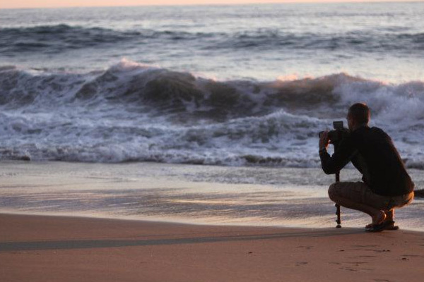A person squats on a sandy beach near the shoreline, holding a camera and tripod—perhaps capturing their study abroad adventures—as ocean waves roll in and soft light illuminates the scene.