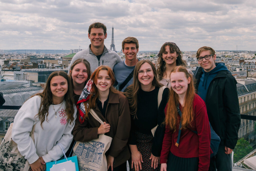 A group of nine young adults pose and smile on a rooftop with a cityscape and the Eiffel Tower in the background under a cloudy sky.