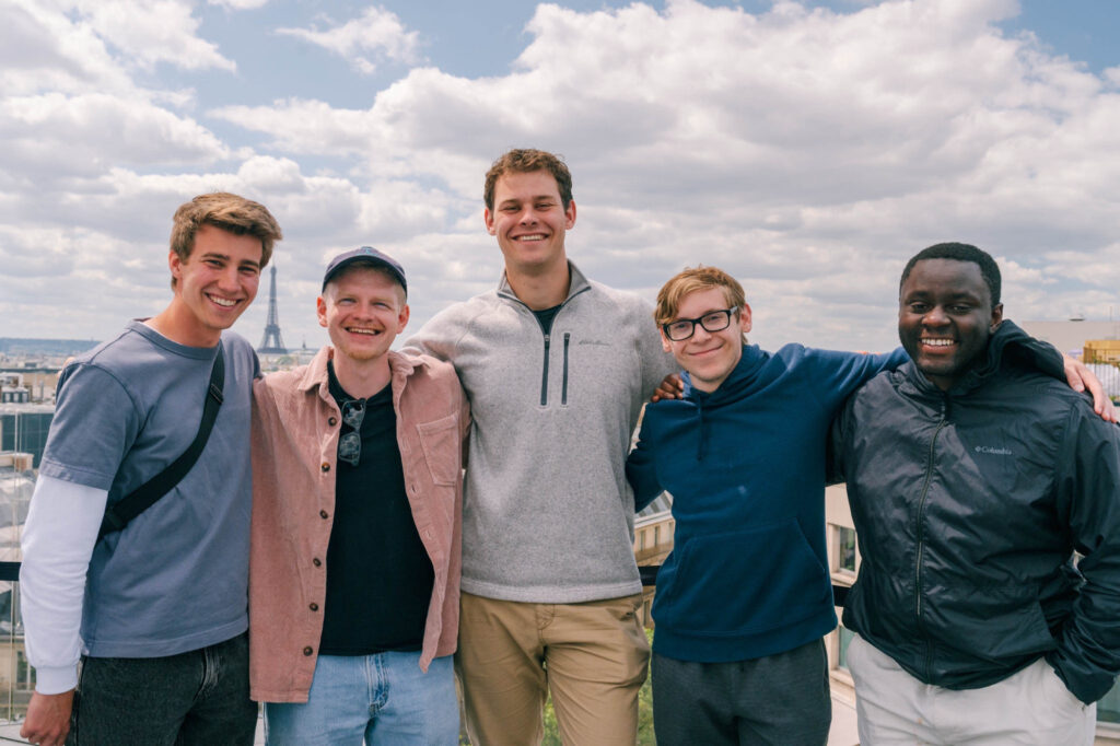 Five young men pose together outdoors on a rooftop with a cityscape in the background. They are smiling and standing close under a partly cloudy sky.