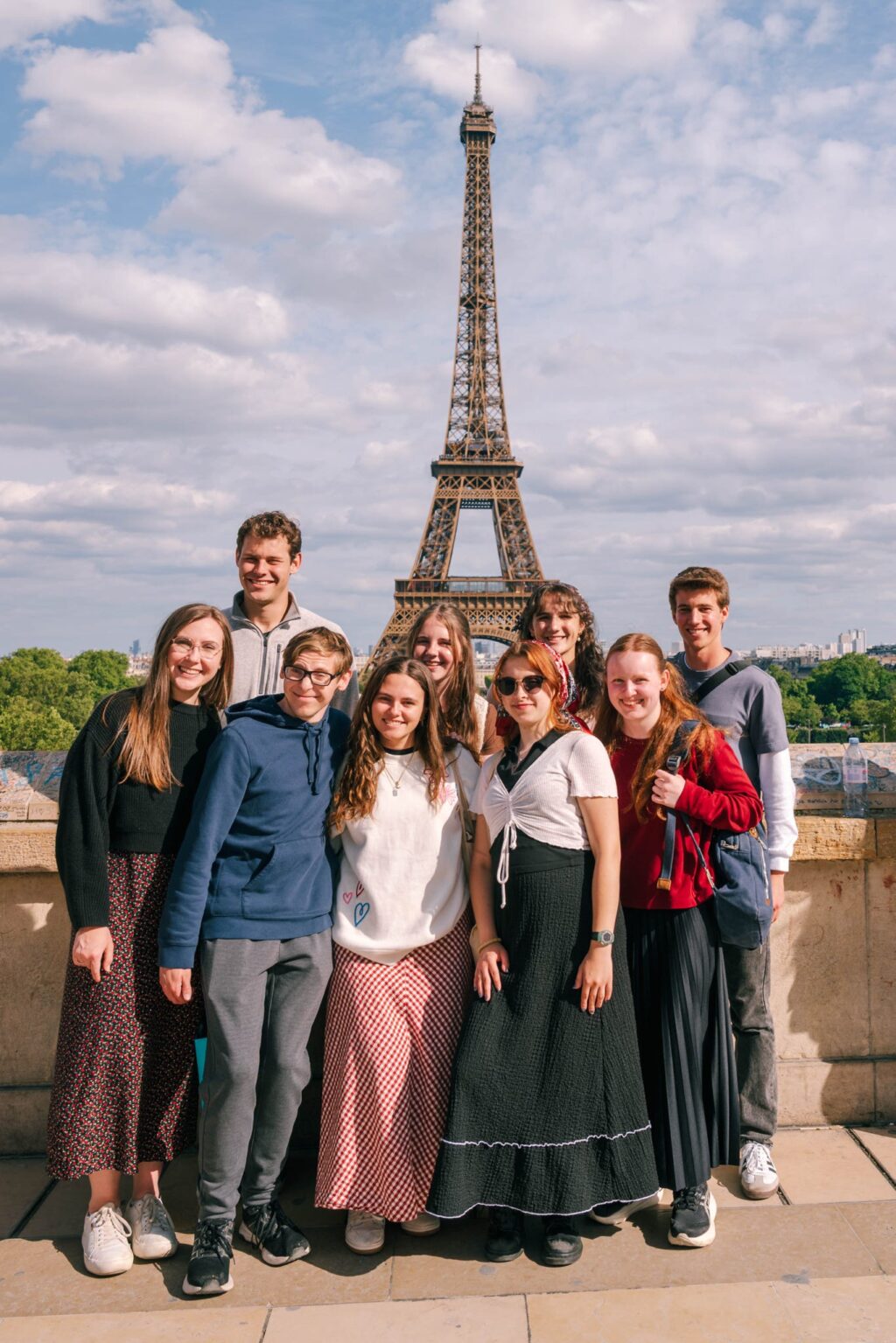 A group of nine young people pose and smile in front of the Eiffel Tower in Paris on a partly cloudy day. They are dressed casually and stand close together, with trees and the city visible in the background.