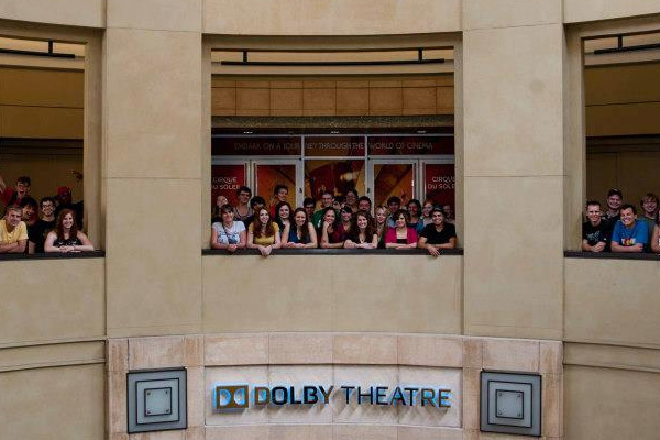 A group of people, possibly Study Abroad participants, stand behind a railing on a balcony above a sign that reads Dolby Theatre. The setting appears to be indoors, with beige walls and large windows in the background.