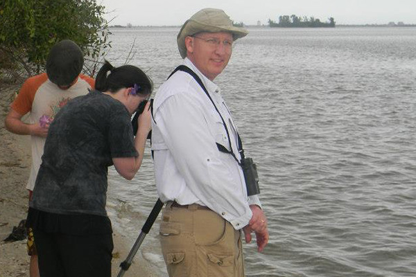Three people stand on a shoreline. The person in front, wearing a hat and binoculars, faces the camera. The others, perhaps study abroad students in casual clothes, look down and use a camera or binoculars, facing away from the water.