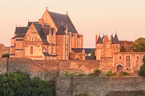 A large stone castle with multiple gabled roofs and towers sits behind a stone wall, illuminated by warm sunlight. Perfect for those dreaming to study abroad, the scene is framed by trees and greenery in the foreground.