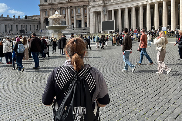 A person with a backpack stands in a large cobblestone square filled with people, embodying the spirit of study abroad, with a stone fountain and a historic building featuring columns and statues in the background.