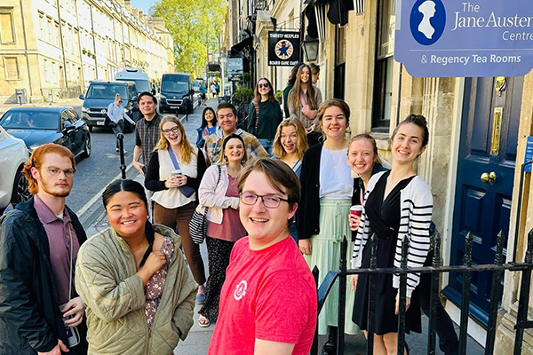 A group of people stand smiling on a city sidewalk outside The Jane Austen Centre and Regency Tea Rooms, sharing drinks. Their Study Abroad adventure brings lively energy to the street, with buildings, parked cars, and a street sign in the background.