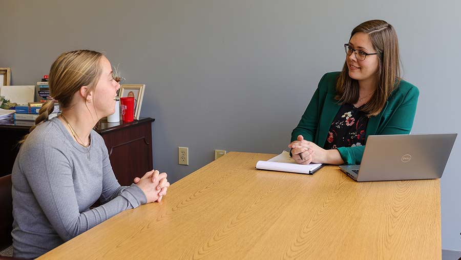 Two women sit across from each other at a wooden table in an office. One is taking notes on résumé writing with a notepad and pen, her laptop open in front of her, while the other woman sits with her hands clasped.