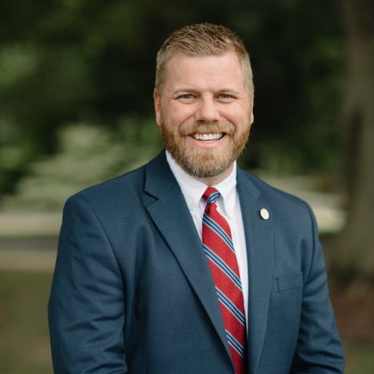 Stephen Presley, a man with short blond hair and a beard, is wearing a blue suit, white shirt, and red striped tie. He is smiling and standing outdoors near the chapel, with trees and greenery blurred in the background.