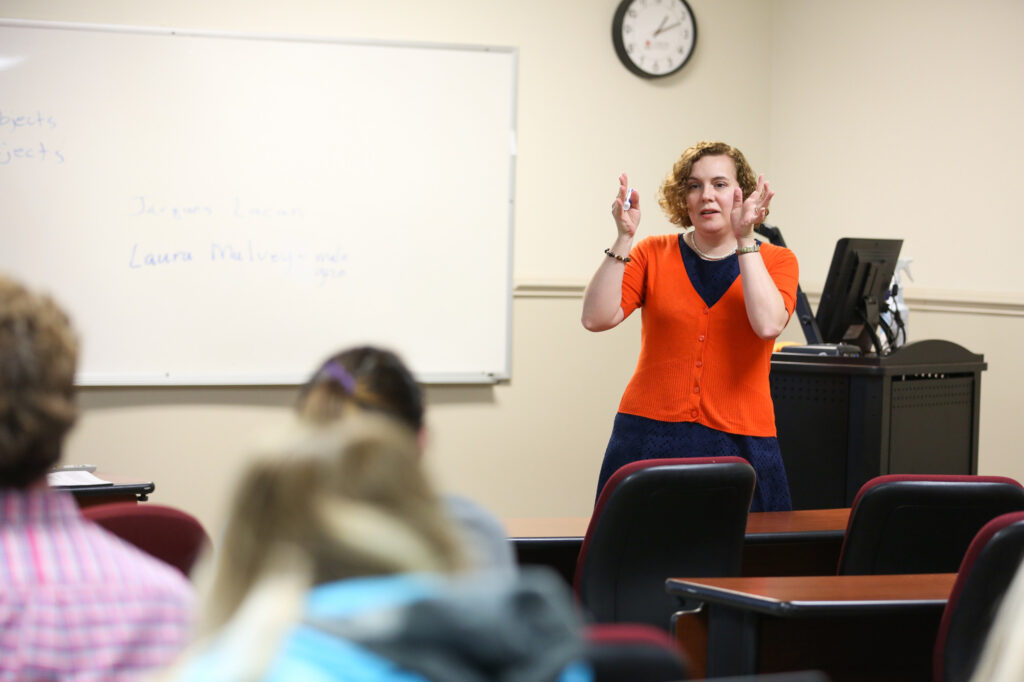 A woman in an orange cardigan stands at the front of a classroom, gesturing with her hands while speaking. Several students face her, and a whiteboard with writing and a clock are visible behind her.